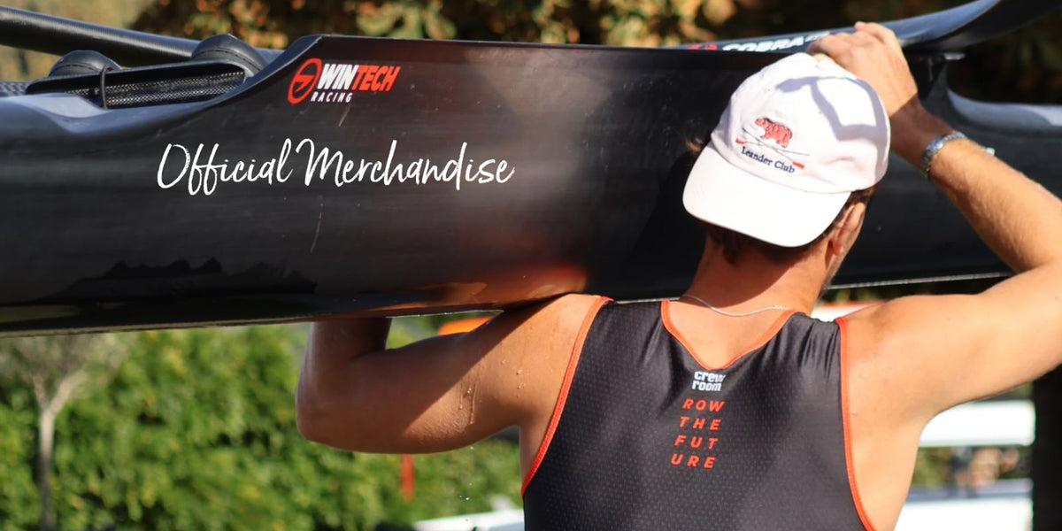 A person outdoors carrying a black rowing boat on their shoulder, wearing a white cap and a black athletic rowing suit with orange trim. The boat has 'WinTech' and 'Official Merchandise' branding.